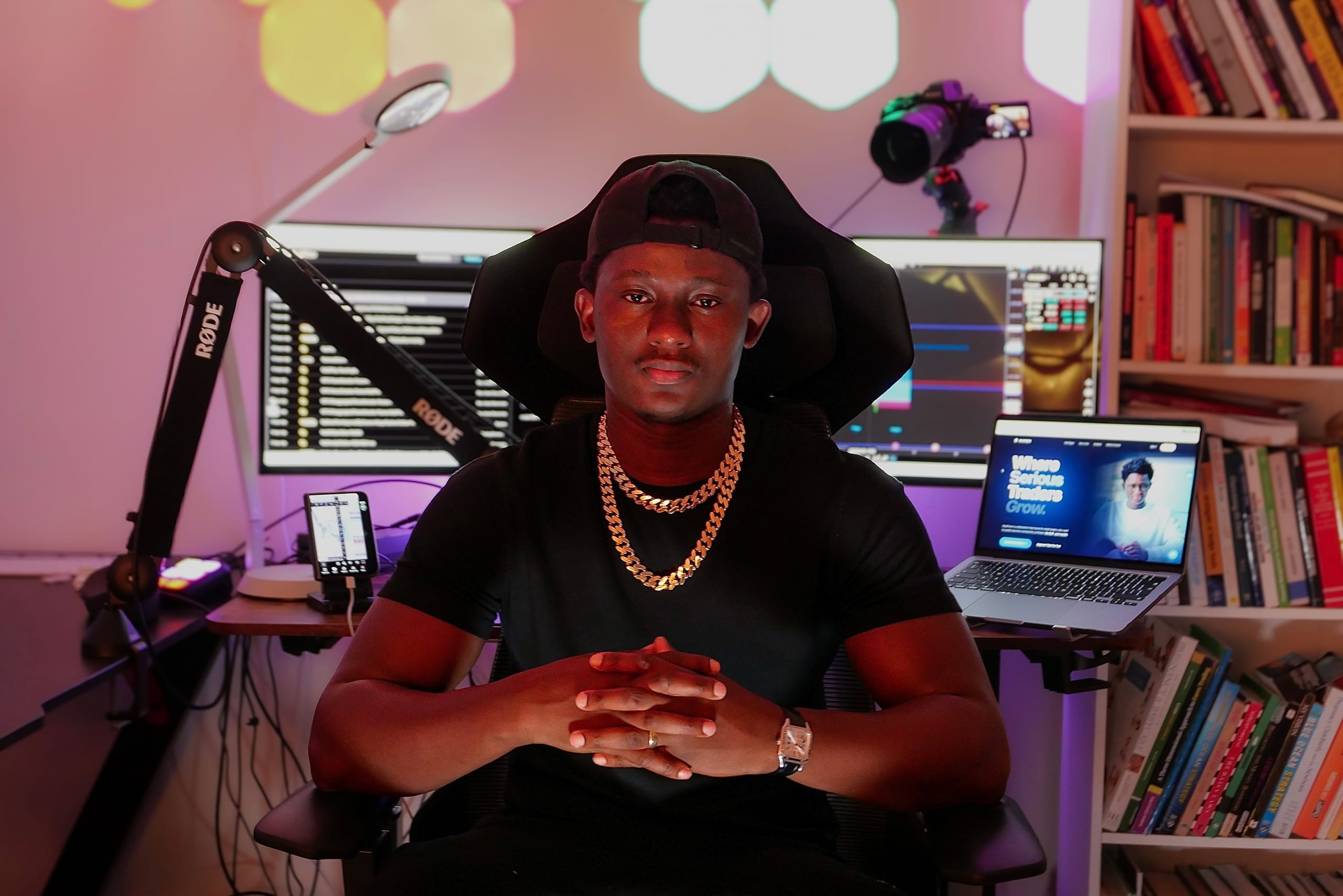 KojoForex at his trading desk under neon light, gold chains, hands clasped